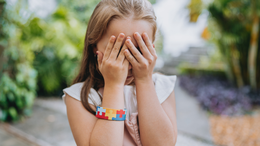 Niña con brazalete que lleva el símbolo autista, se tapa la cara con las manos.
